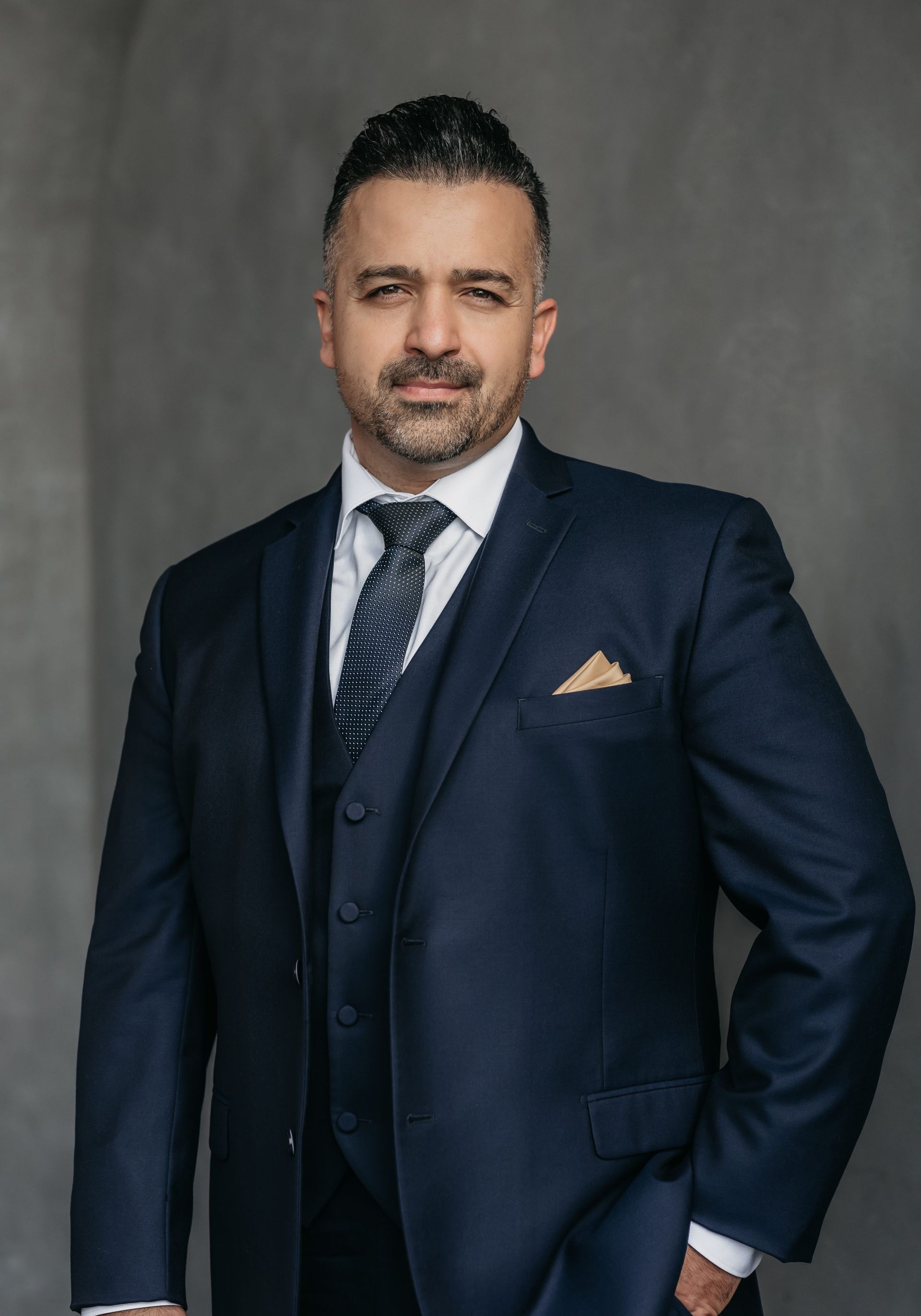 A man in a dark suit and tie stands against a grey background, looking confidently at the camera with one hand in his pocket.