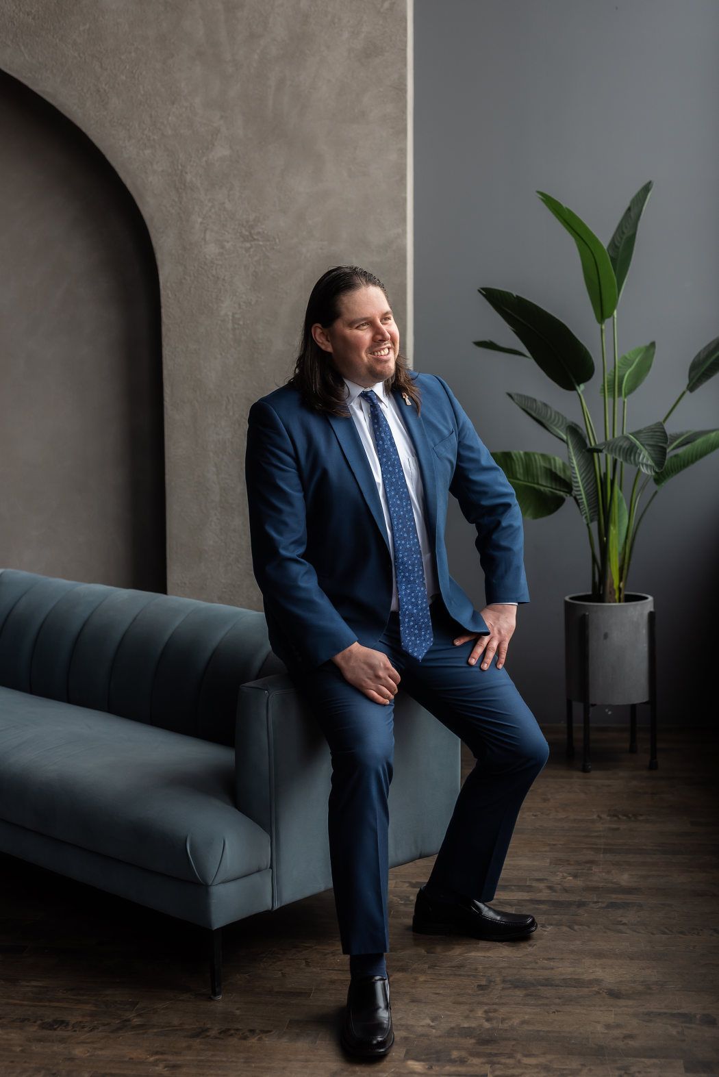 A well-dressed gentleman in a blue suit rests his arm on the back of a beige couch, with a bright smile on his face.