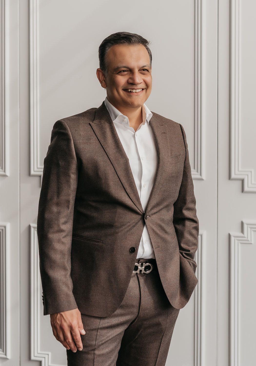 A man wearing a suit and tie stands against a clean white wall, exuding professionalism.