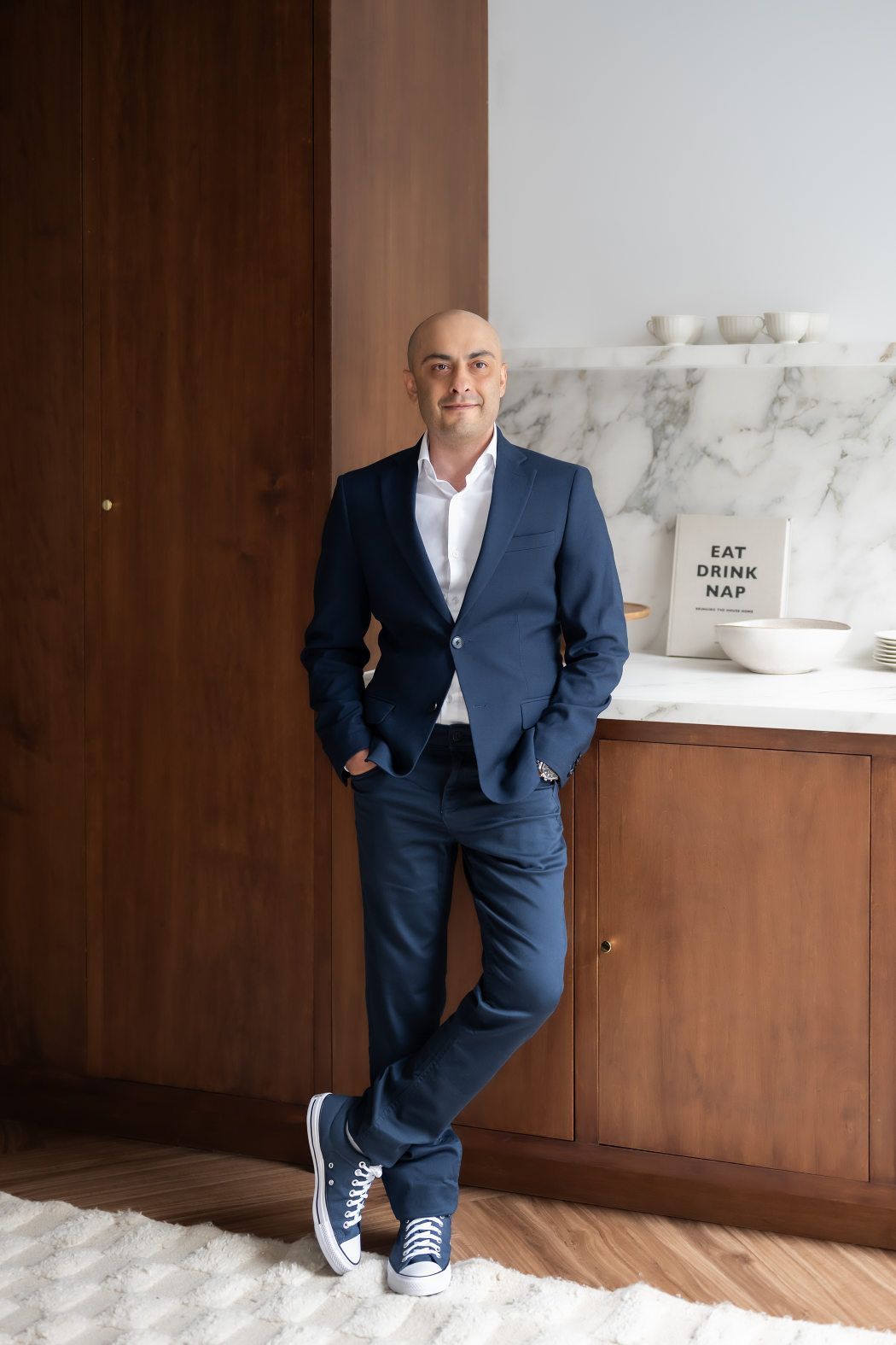 A well-dressed man is standing in front of a cabinet with a light smile on his face, and both his hands are in his pockets.