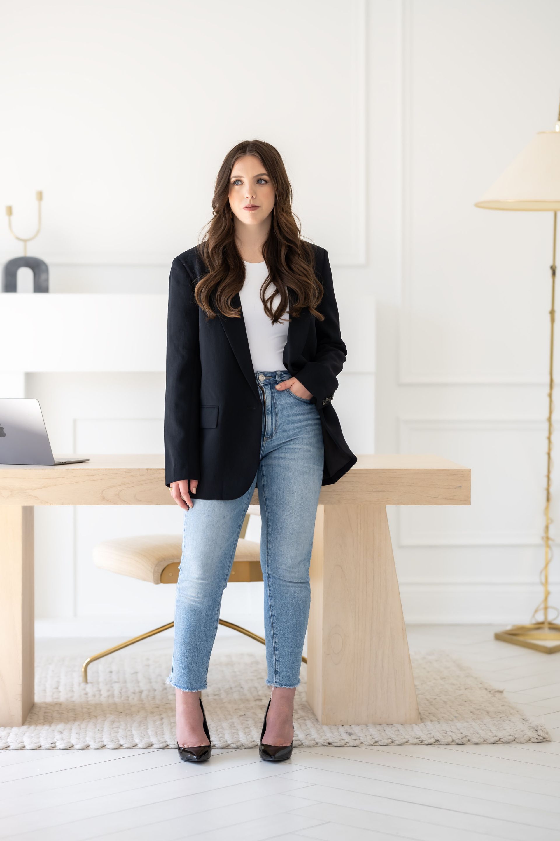 A woman in a black blazer and jeans is standing in front of a desk.