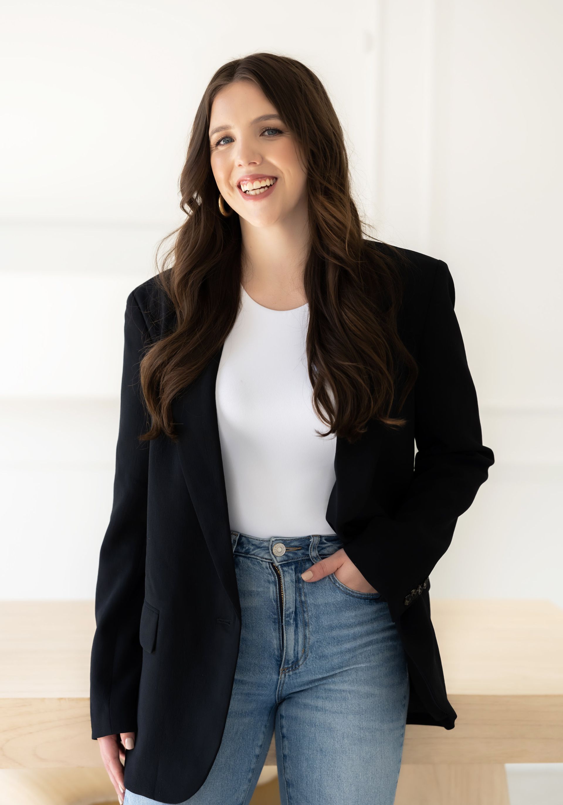 Close-up of a smiling woman with cascading curly hair, dressed in jeans, a dark jacket, and white top, hand casually in her front pocket, showcased in personal branding pictures.