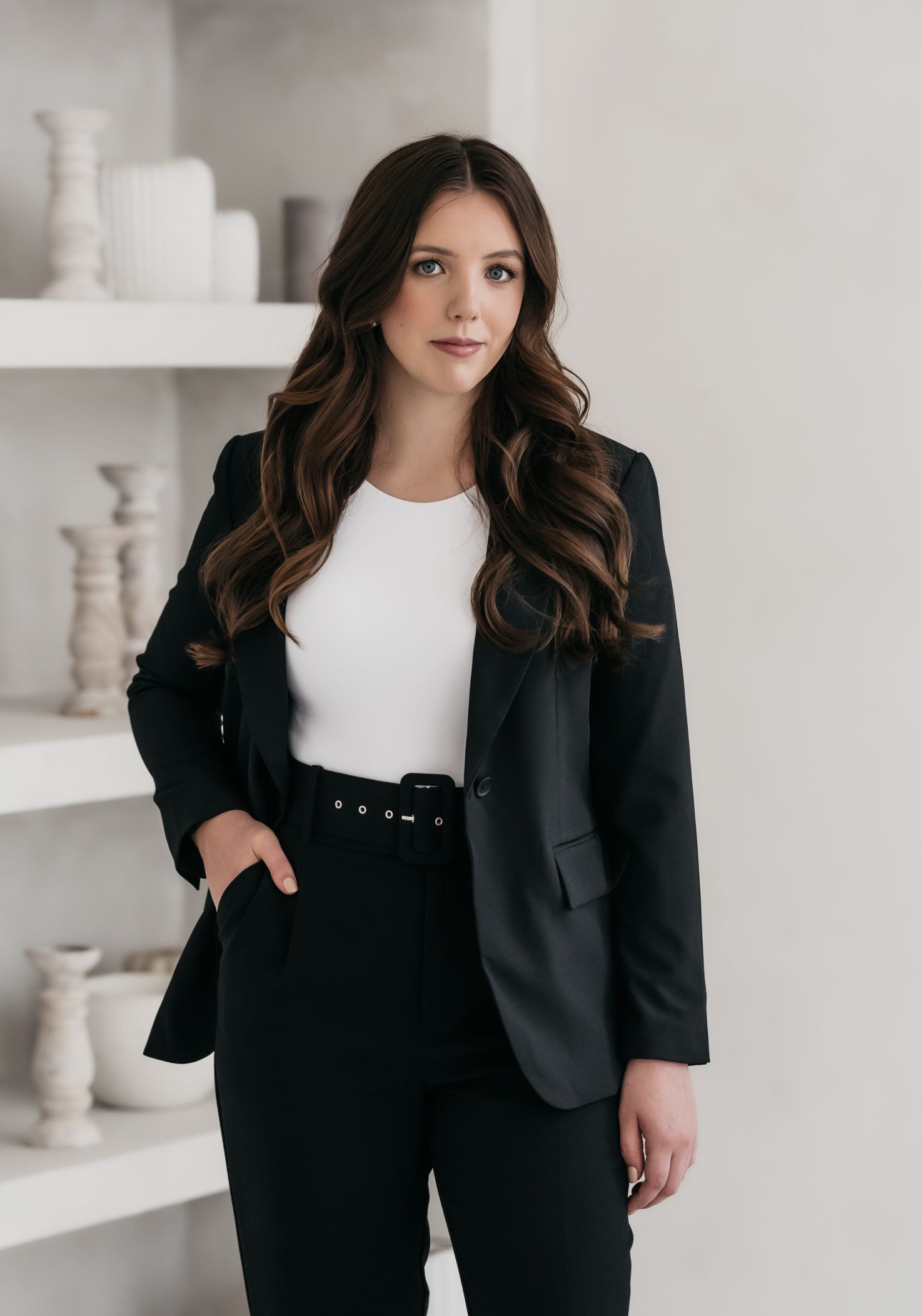 A woman in a black suit and white shirt is standing in front of a bookshelf.
