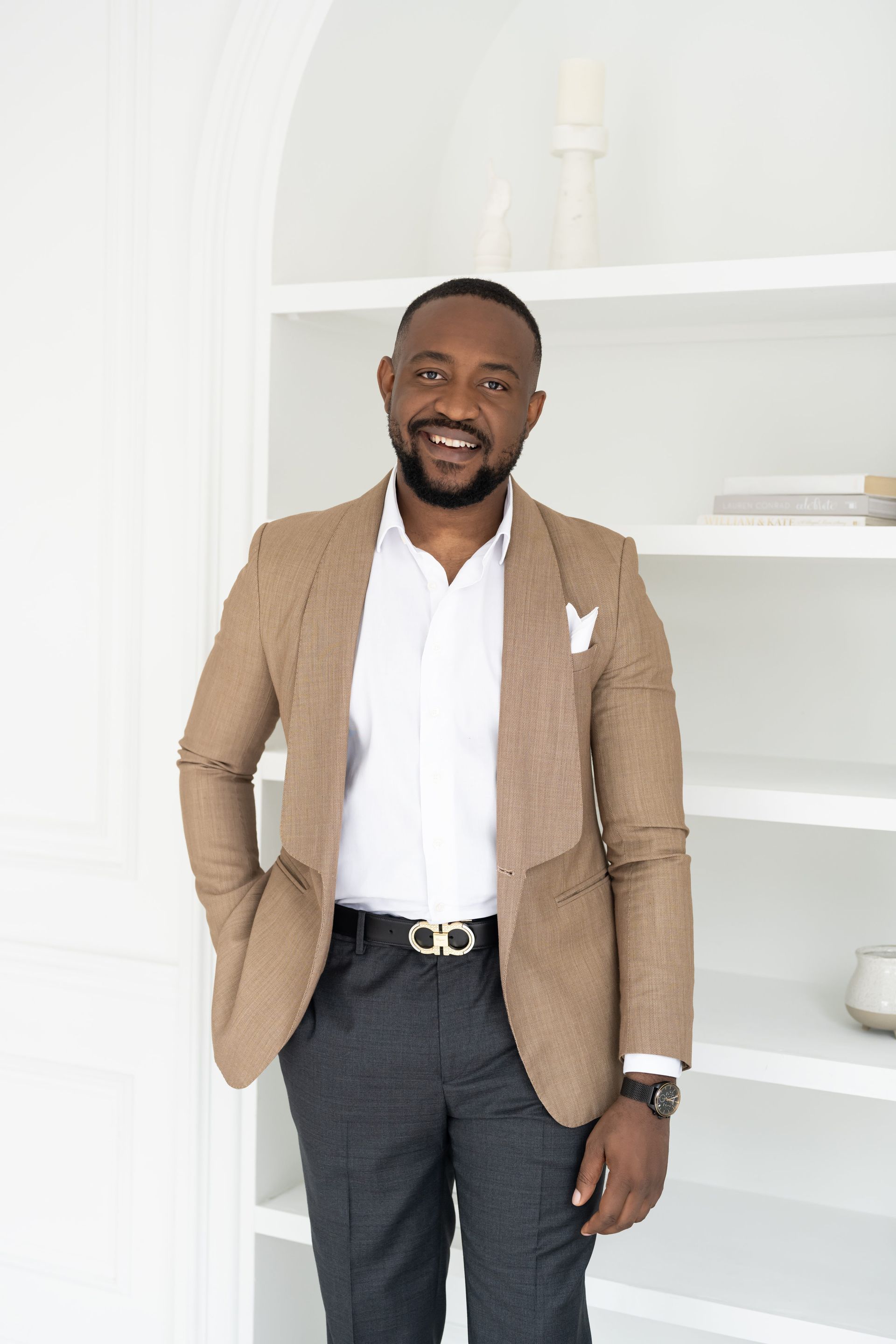 A man stands in front of a bookshelf, in a suit, with his arms at his sides. He has a calm, confident demeanor, and he smiles at the camera.