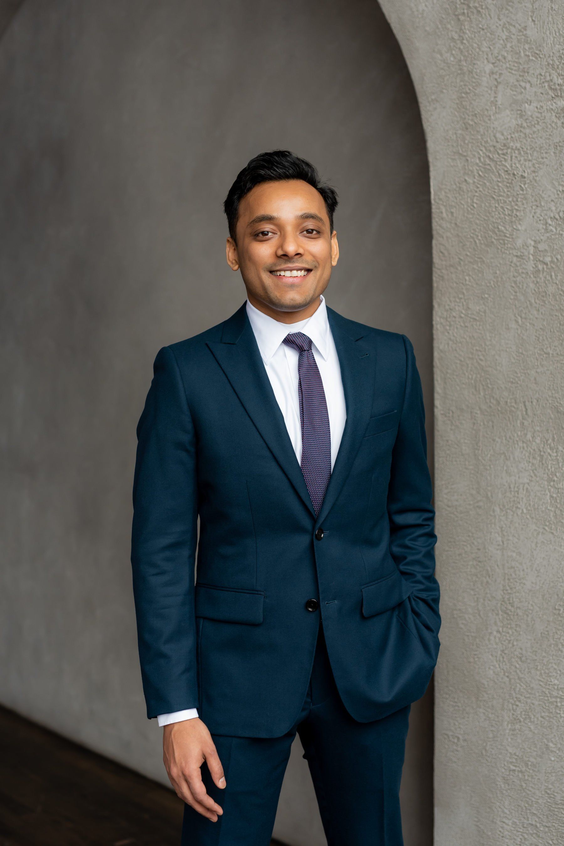 A man with slicked back black hair wearing a navy blue suit posing in a hallway