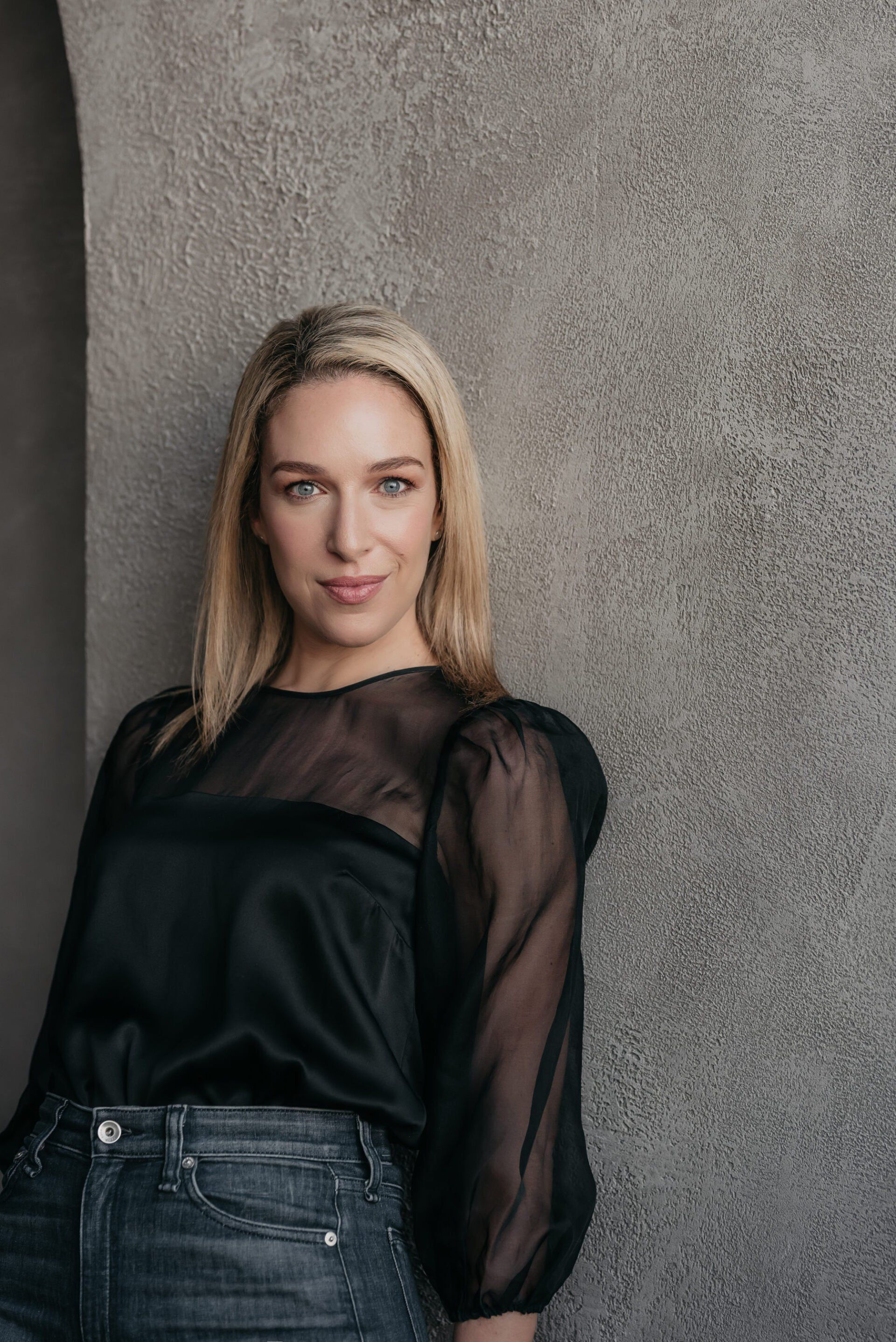 A woman wearing a black mesh top and dark blue jeans leaning on a lime wash wall