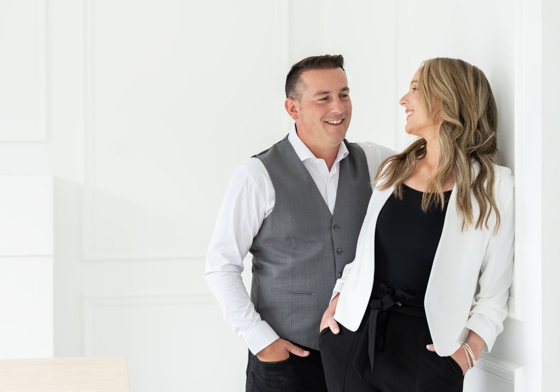 A cheerful couple in semi-formal attire sharing a moment of laughter, with the man in a vest and dress shirt looking at the woman in a white blazer and black top, set against an elegant white paneled background.