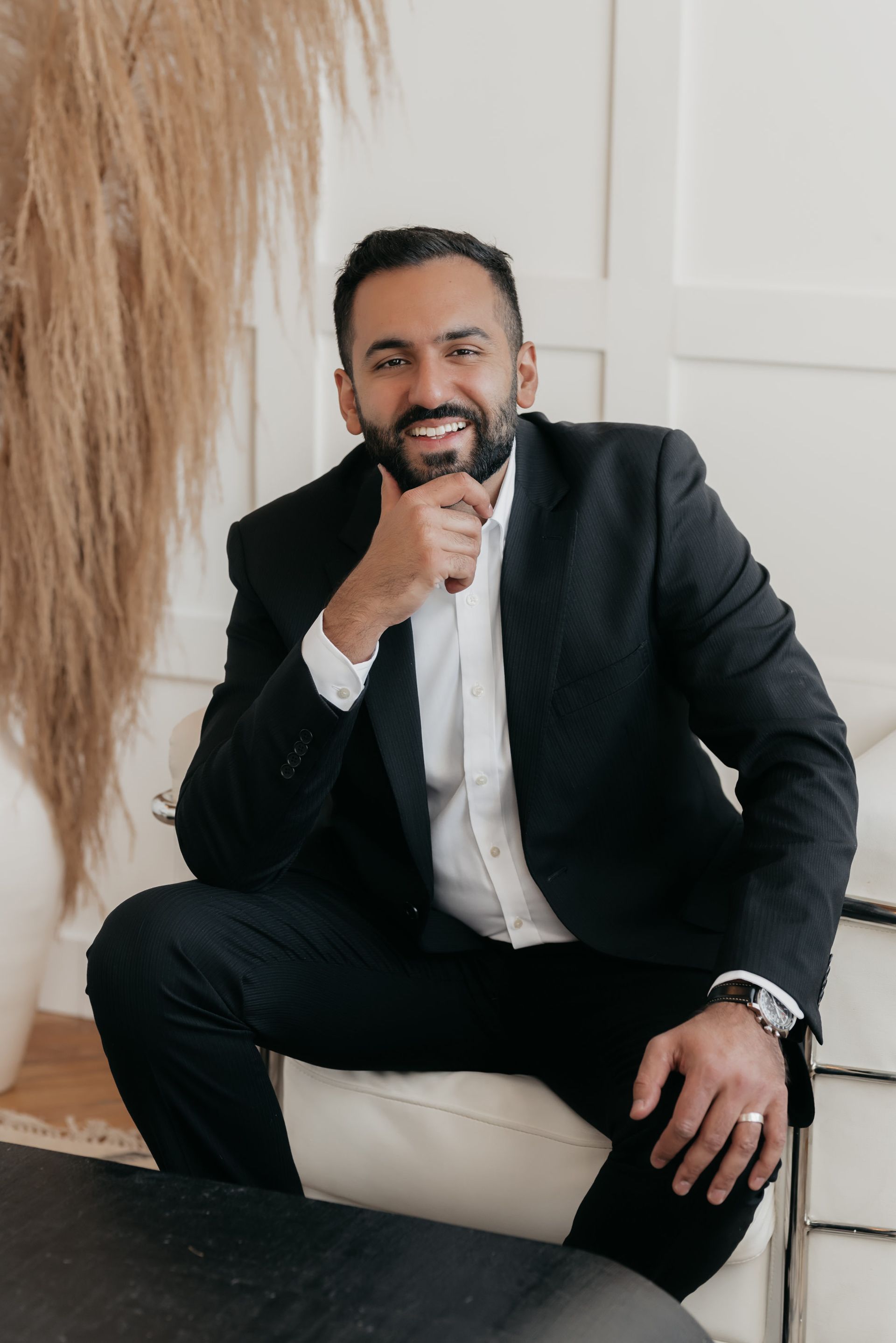 Bearded man in a dark suit and open white shirt, smiling and thoughtfully positioned with one hand on chin, against a pristine white room backdrop featuring a vase with a large feathery plume.