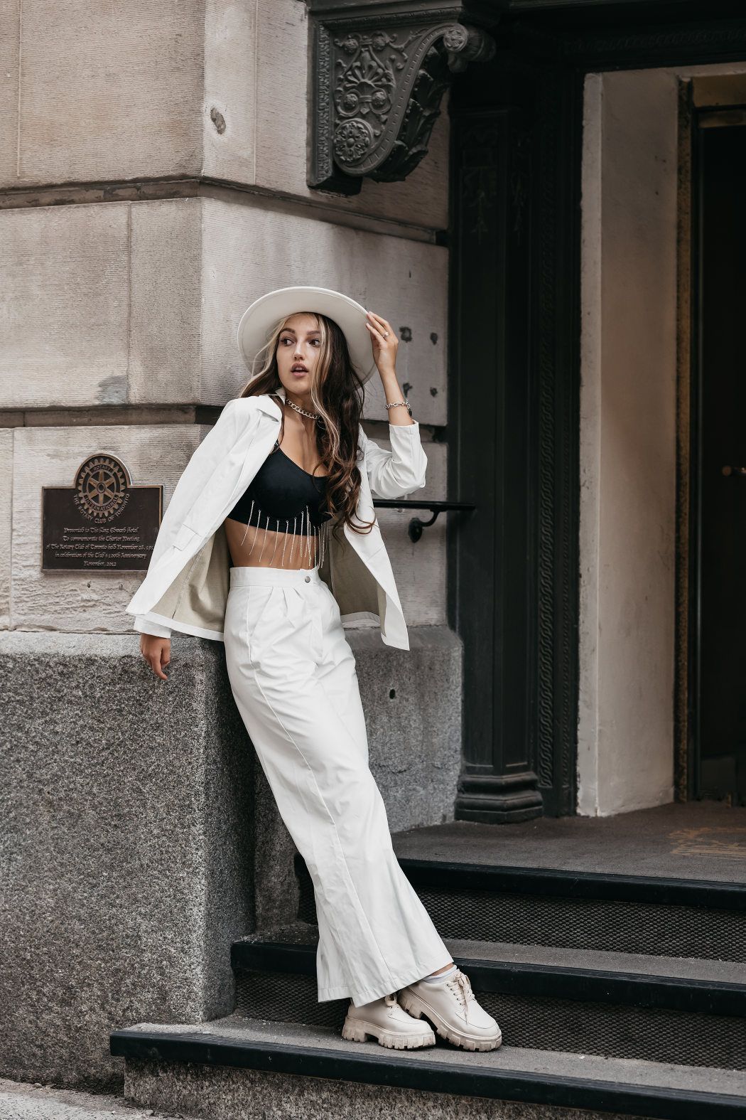 Woman in wide-legged pants (culottes), white jacket, and black crop top with tassels, anchoring a large-brimmed white hat against a breeze, positioned on steps of an aged commercial building, casting a sidelong glance
