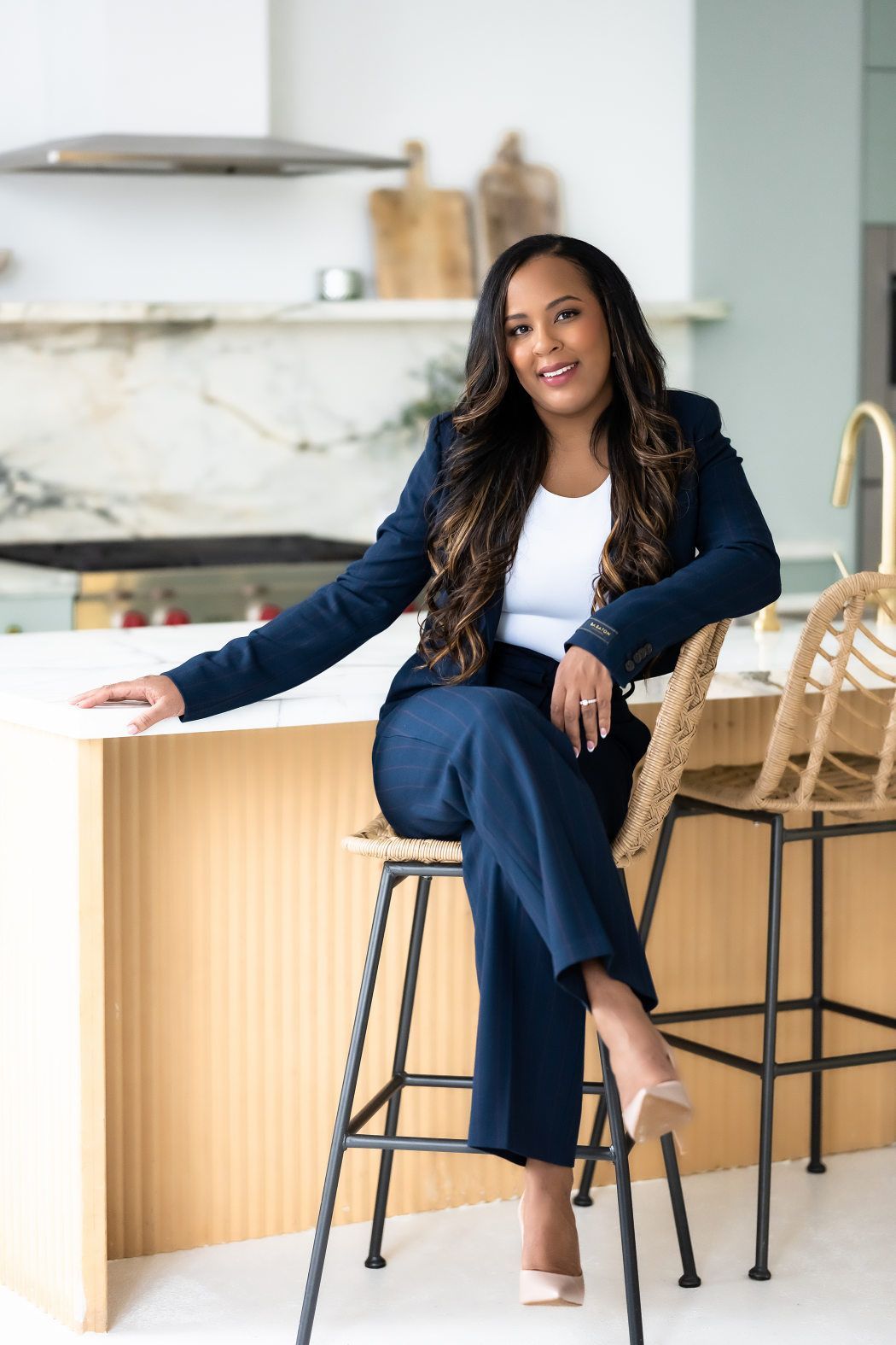 A woman in a suit sitting on a stool in a kitchen, posing for headshots.