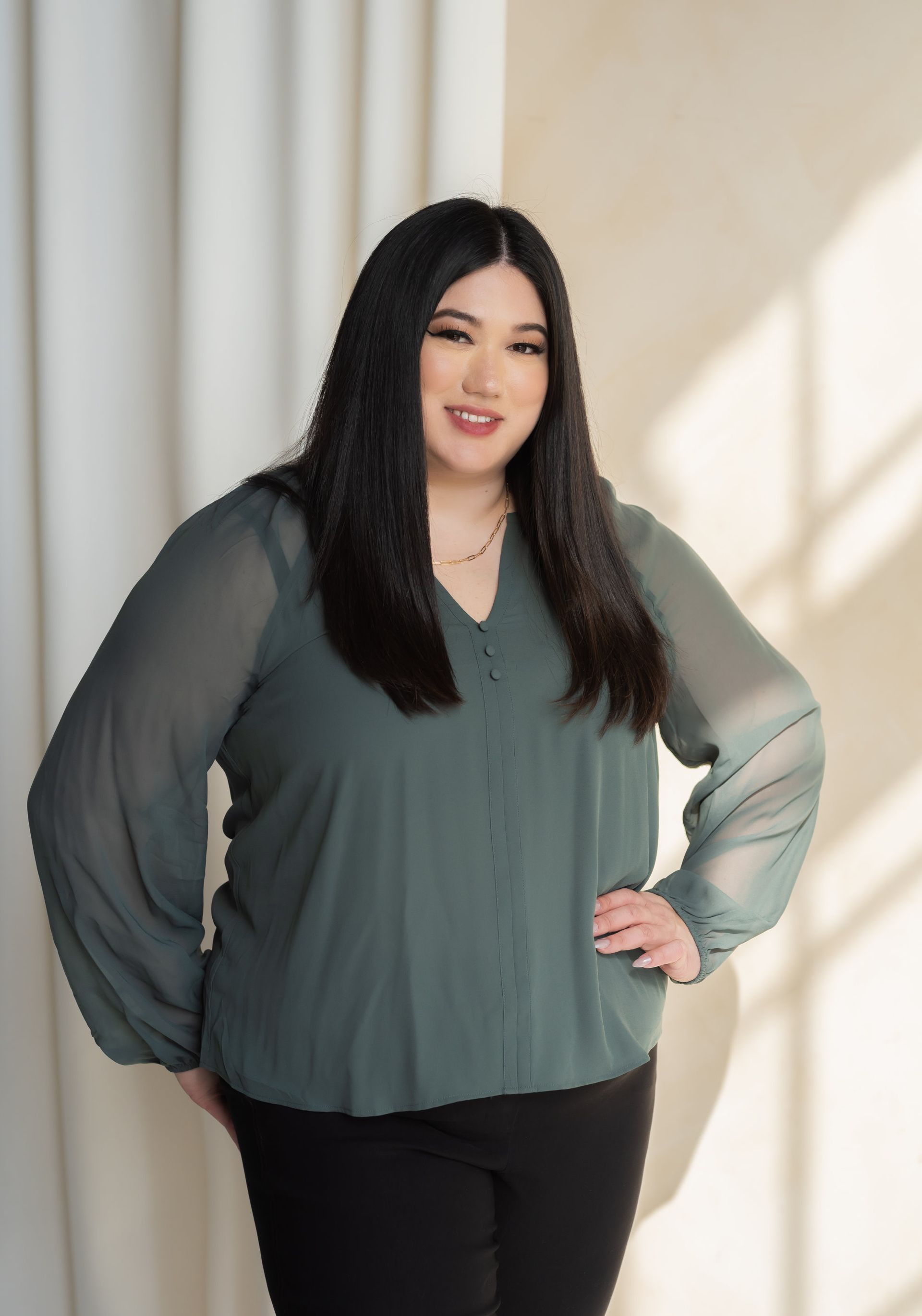 A person stands confidently in a soft green blouse and black pants, against a backdrop of light and draped fabric.