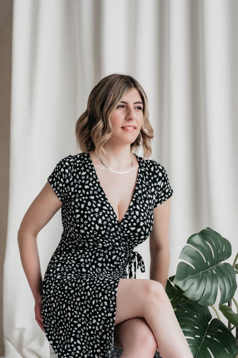 woman in a chic black and white floral dress, looking off-camera with a thoughtful expression, conveying a sense of lifestyle elegance amidst a backdrop of clean curtains and a lush monstera plant.