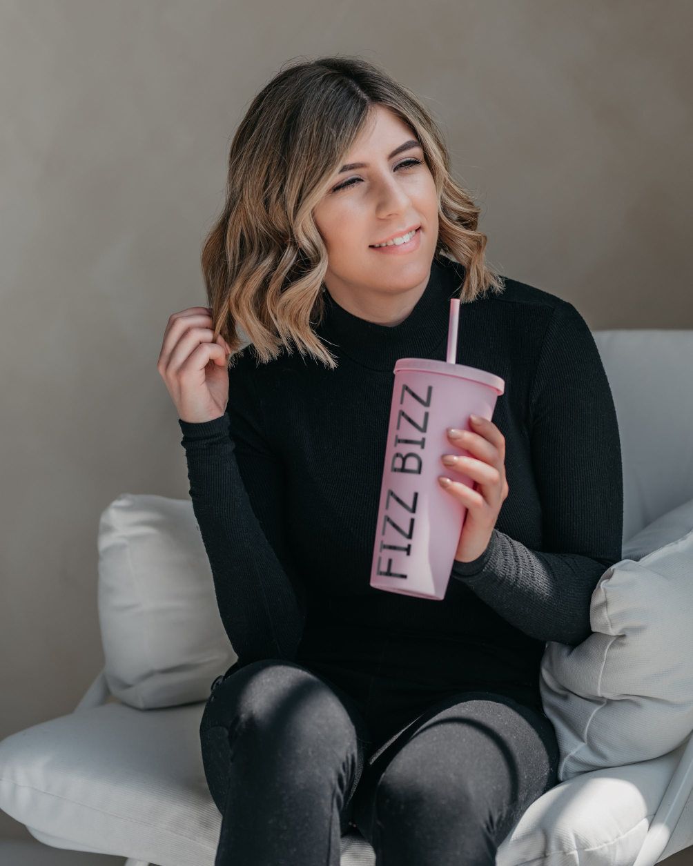 a woman sitting holding a pink drink tumbler, her comfortable black outfit and natural makeup highlighting her personal brand's approachable wellness focus.