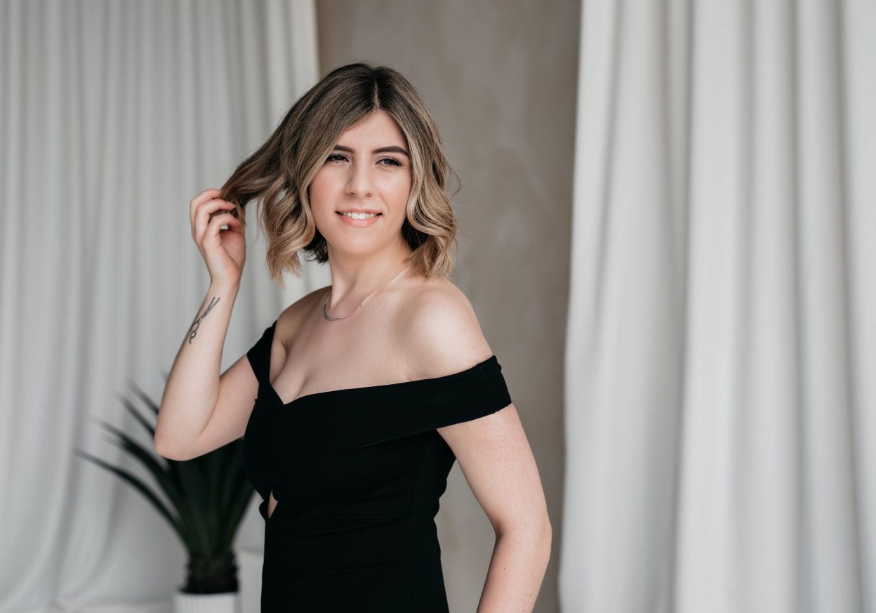 woman in an off-shoulder black dress, her confident stance and engaging smile set against a minimalist background, encapsulating a modern lifestyle and wellness vibe