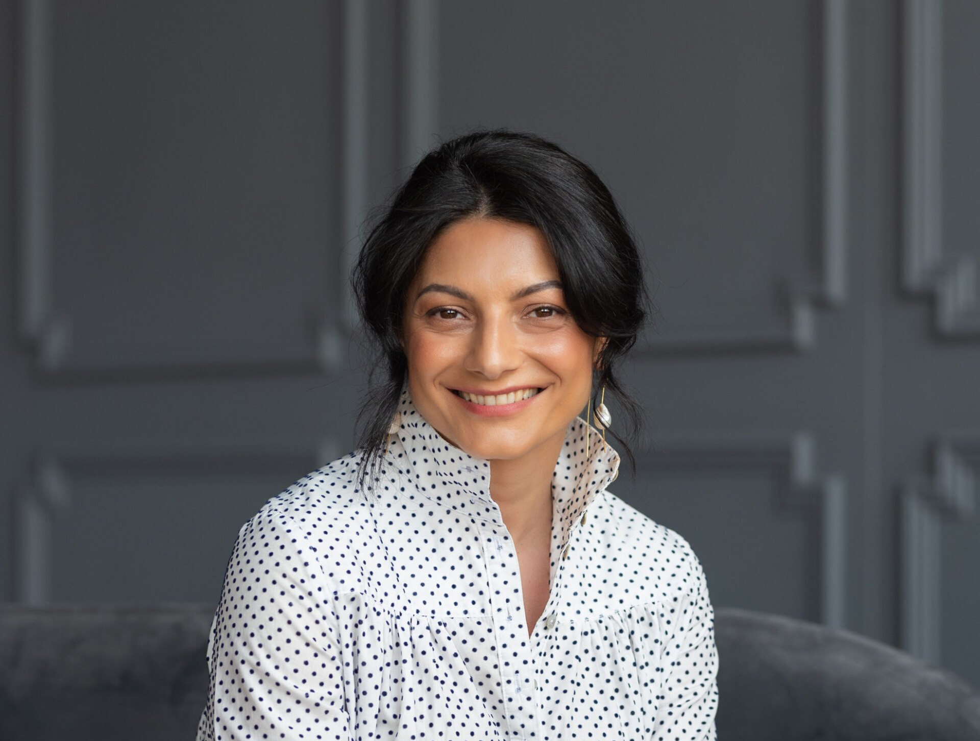A headshot of a woman wearing a polkadot shirt and a grey paneled wall
