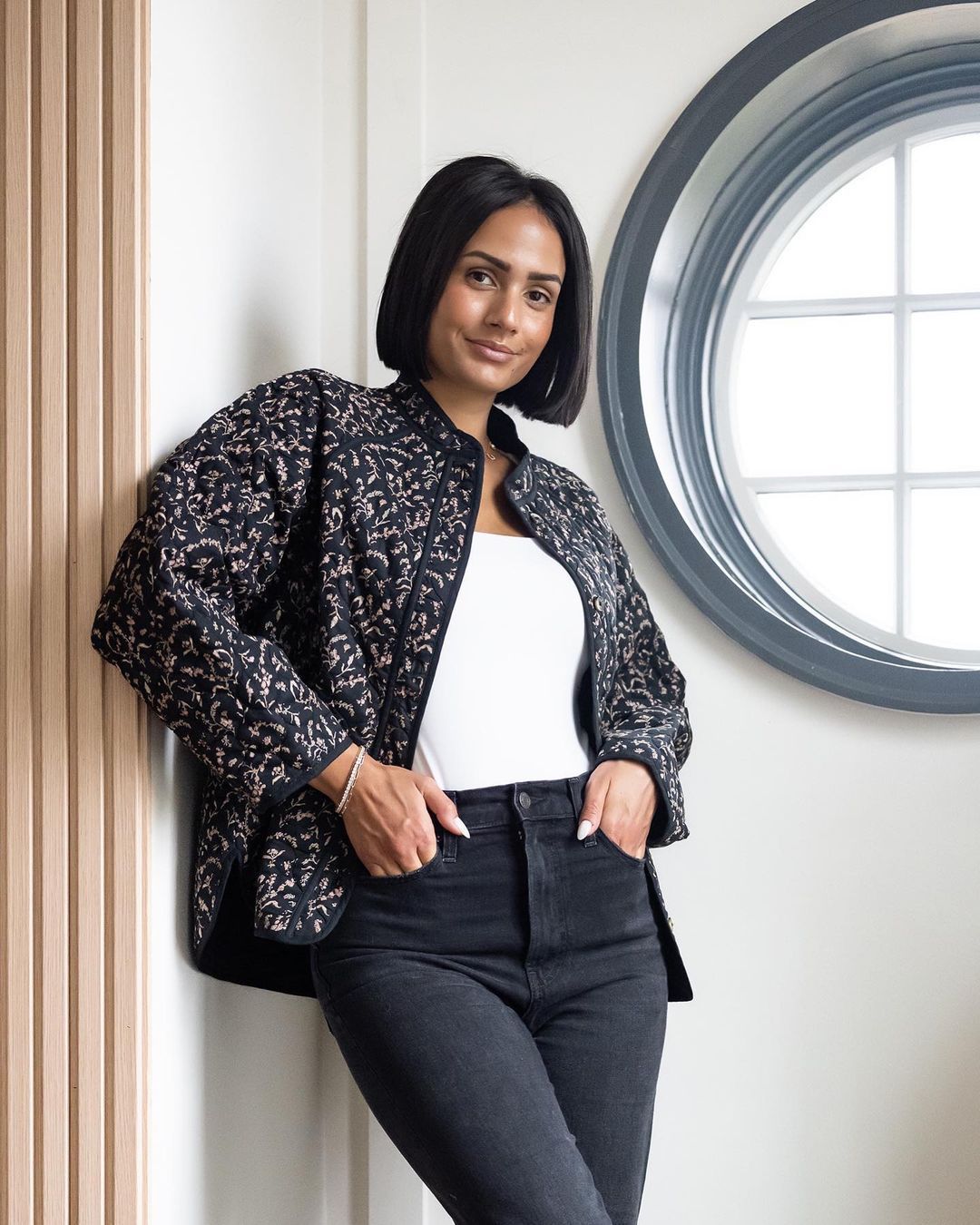 woman with short hair wearing a patterned jacket and white top leaning against the wall