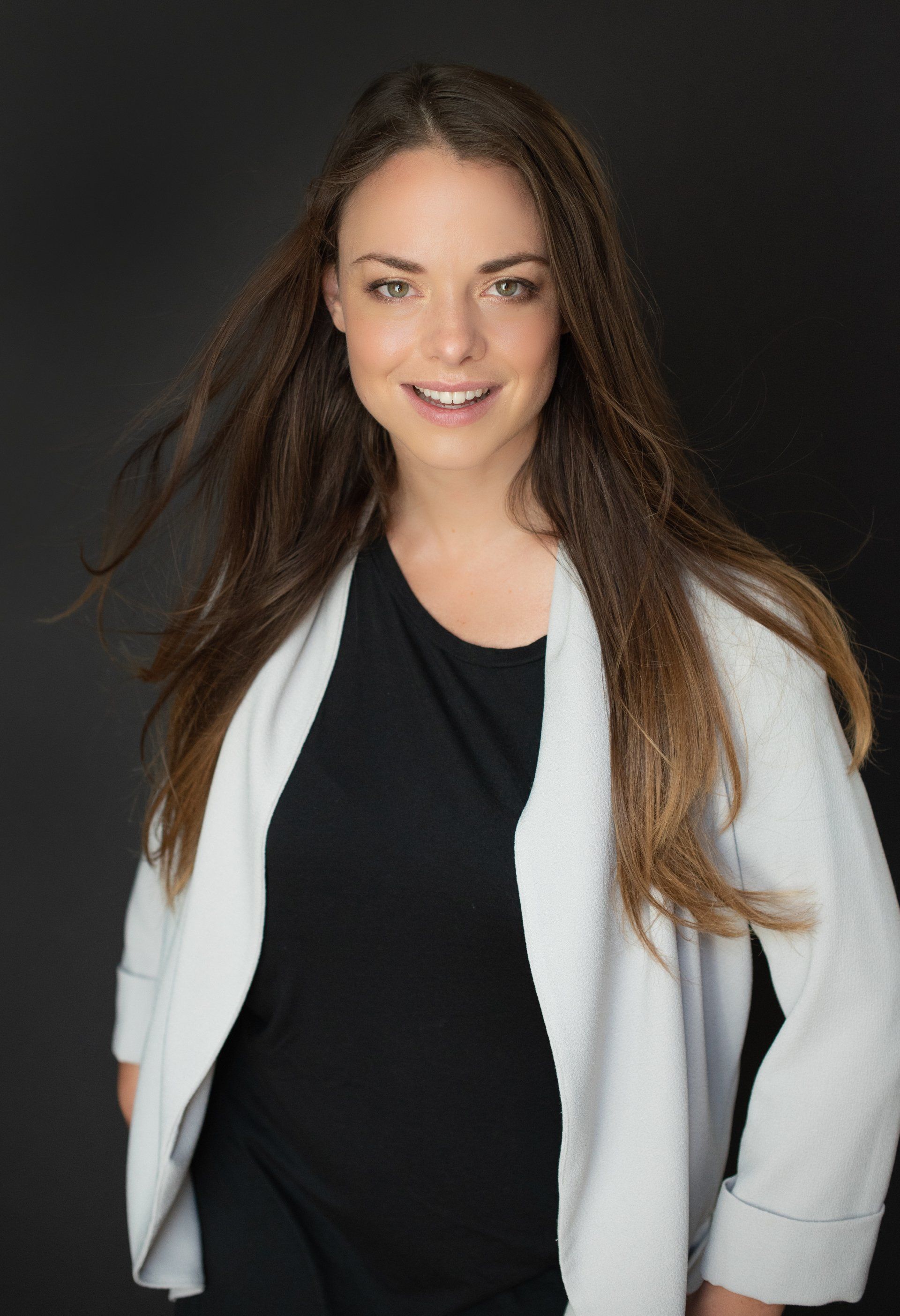 A woman stands in a room with a gray wall, wearing a black shirt and white jacket