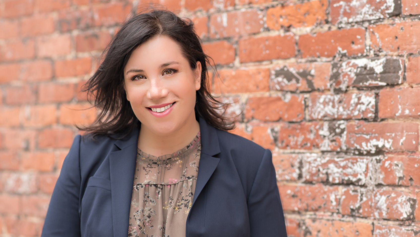 A professional woman in a black blazer standing in front of a brick wall