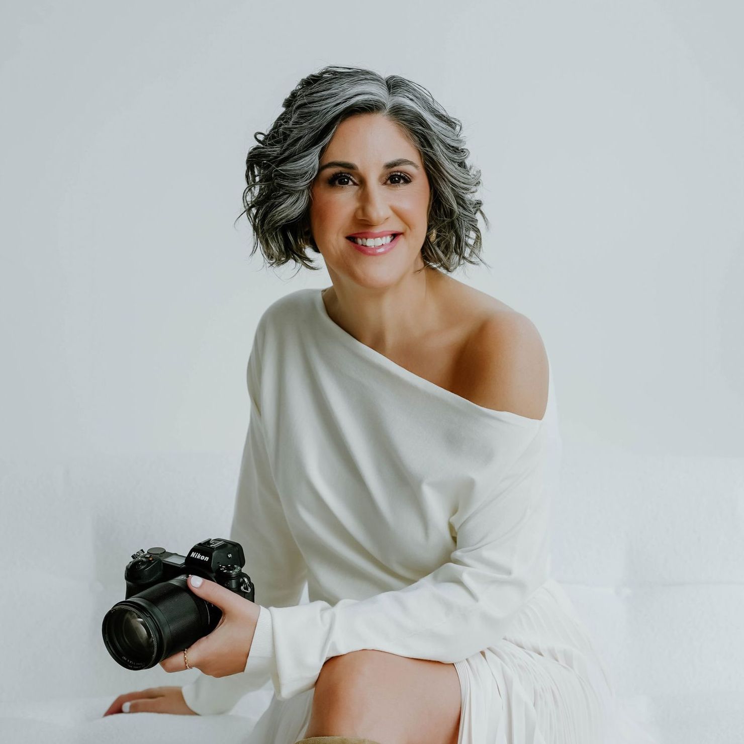 Stacey is in a white dress sitting and  holding a camera.