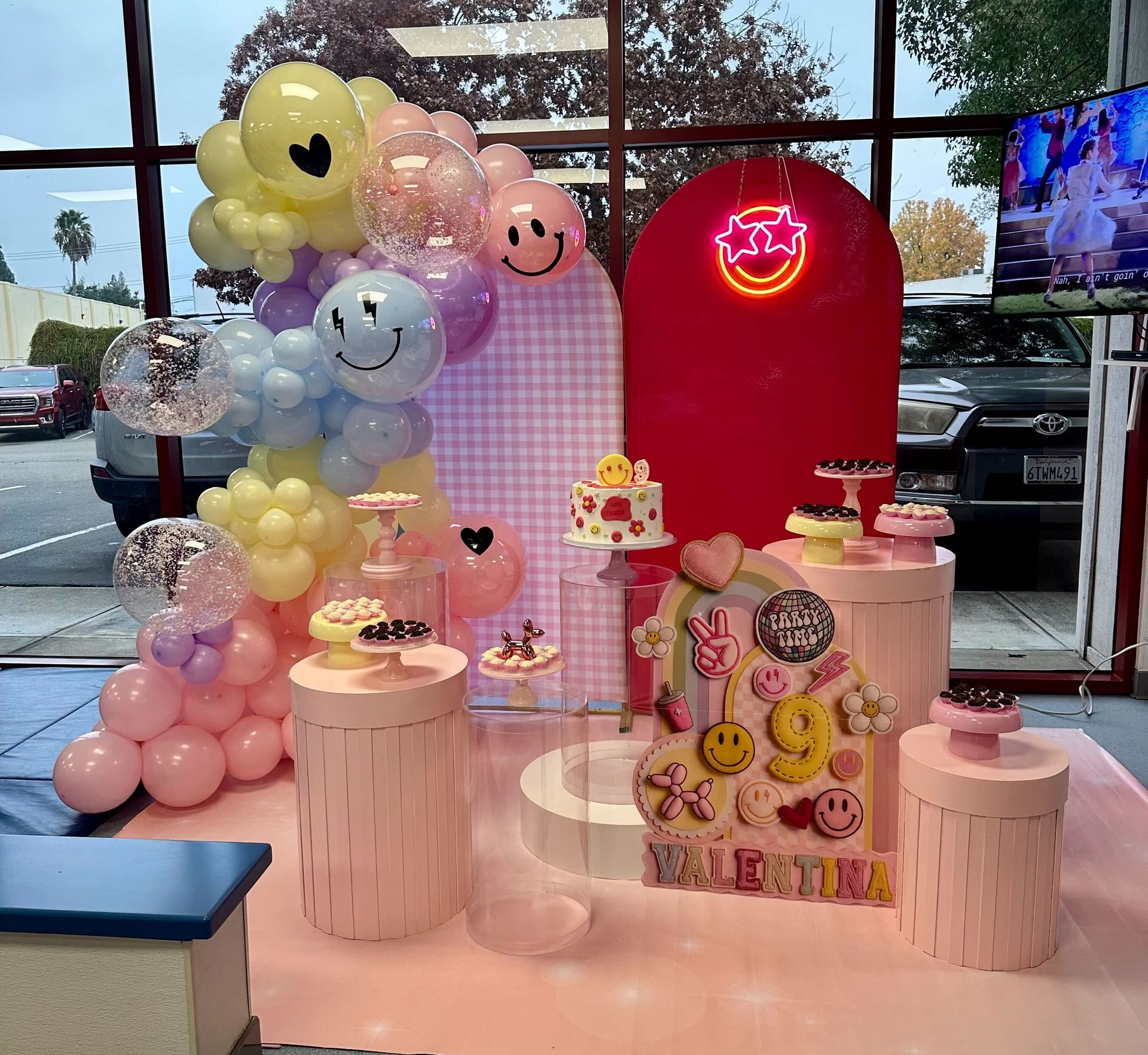 Pastel balloon birthday display with cake stands, a neon sign, and smiling decorative balloons in a shop window