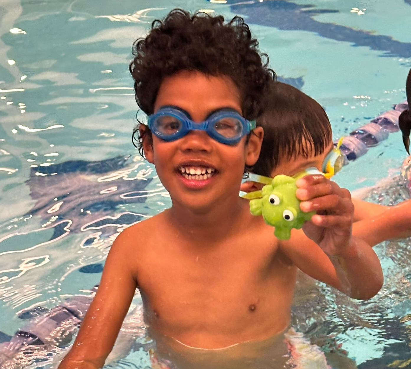 Two children wearing blue goggles smile and splash in a swimming pool