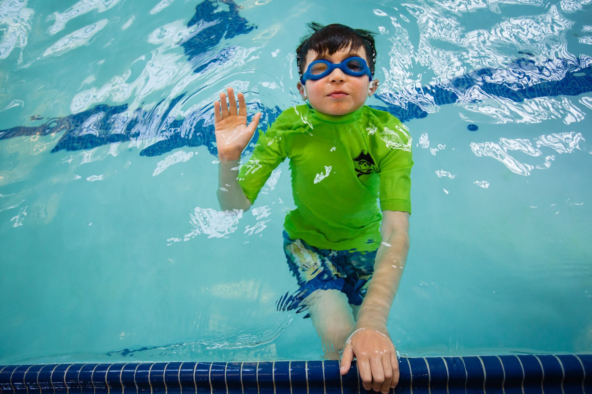 Child swimming underwater in a pool, wearing blue goggles and a green rash guard, holding the pool edge.