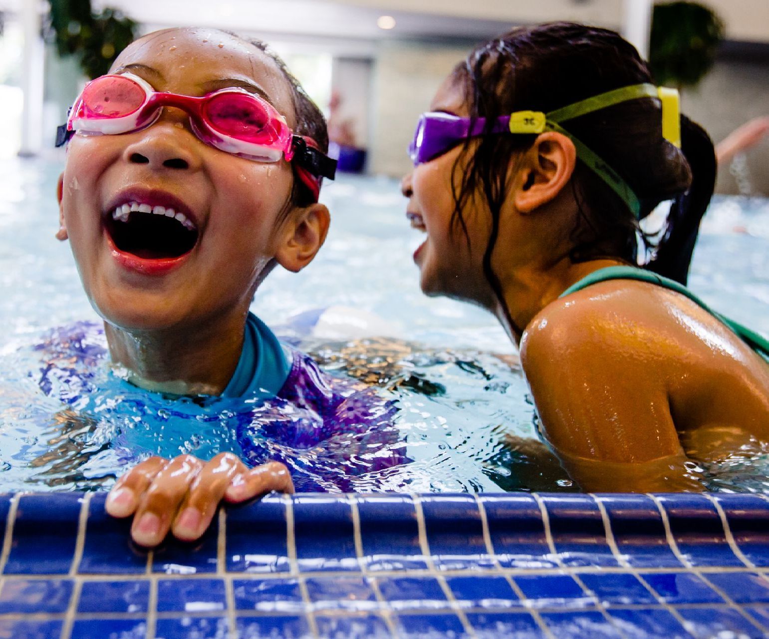Two swimmers at a pool edge, one laughing in pink goggles, one in yellow goggles turning toward them.