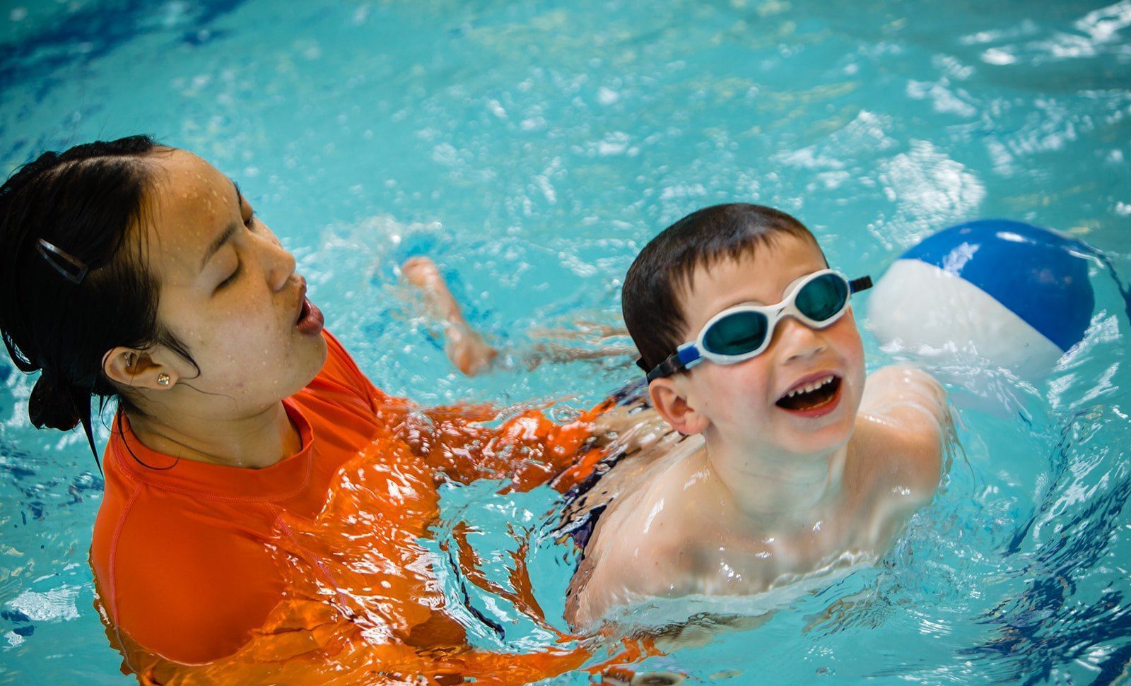 Swimmer in goggles splashing in a pool beside an adult in orange shirt