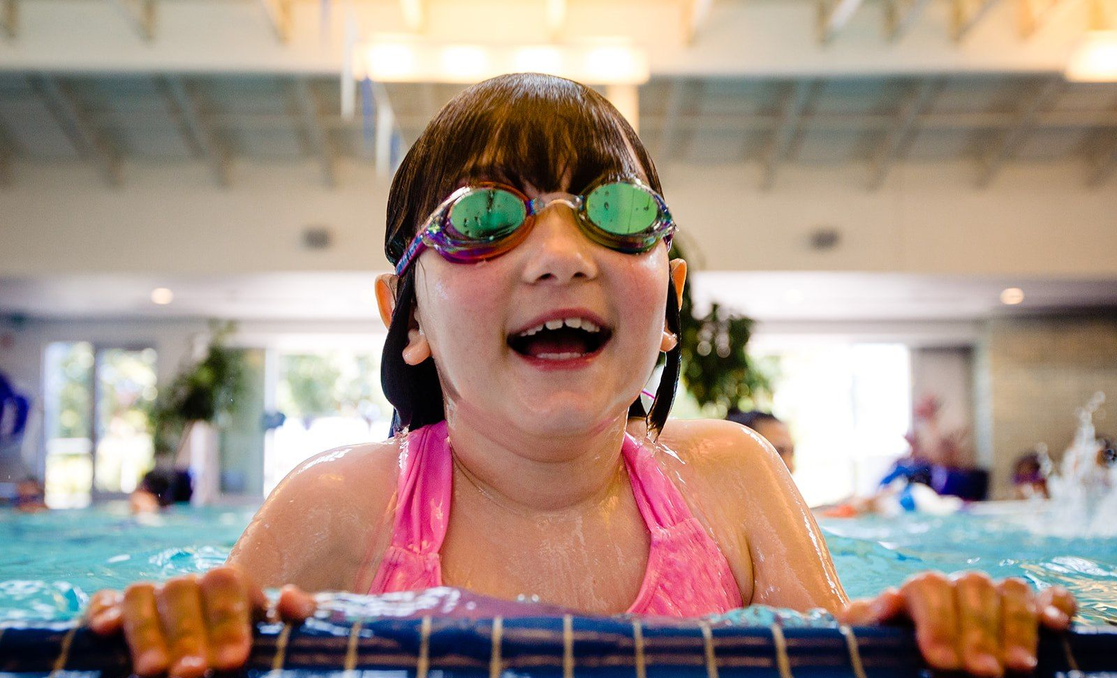 Smiling swimmer in pink goggles and swimsuit holding pool edge indoors
