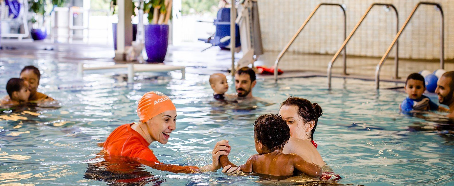 People swimming and playing in an indoor pool, with a coach in an orange shirt guiding them.