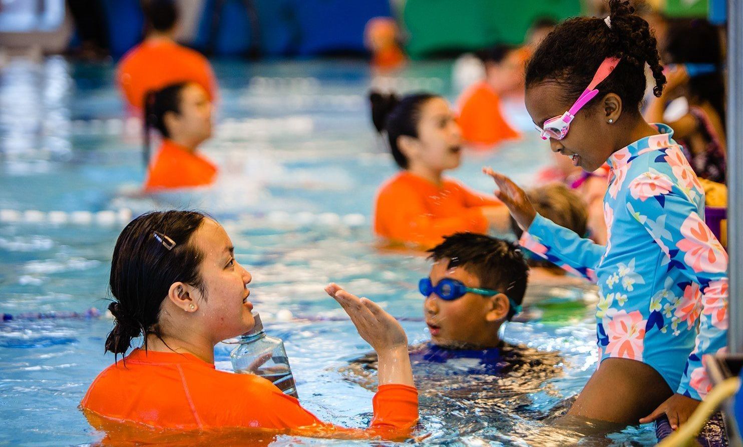 Children and an instructor in a swimming pool, smiling and practicing hand gestures together