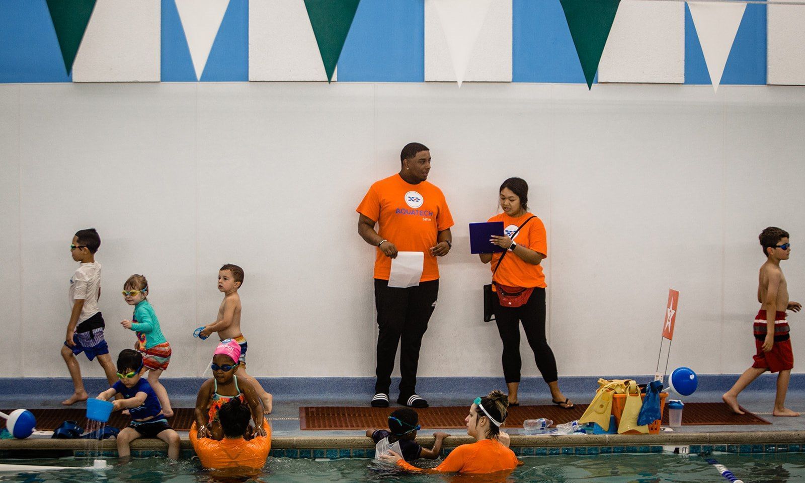 Two lifeguards in orange shirts stand by an indoor pool with swimmers nearby