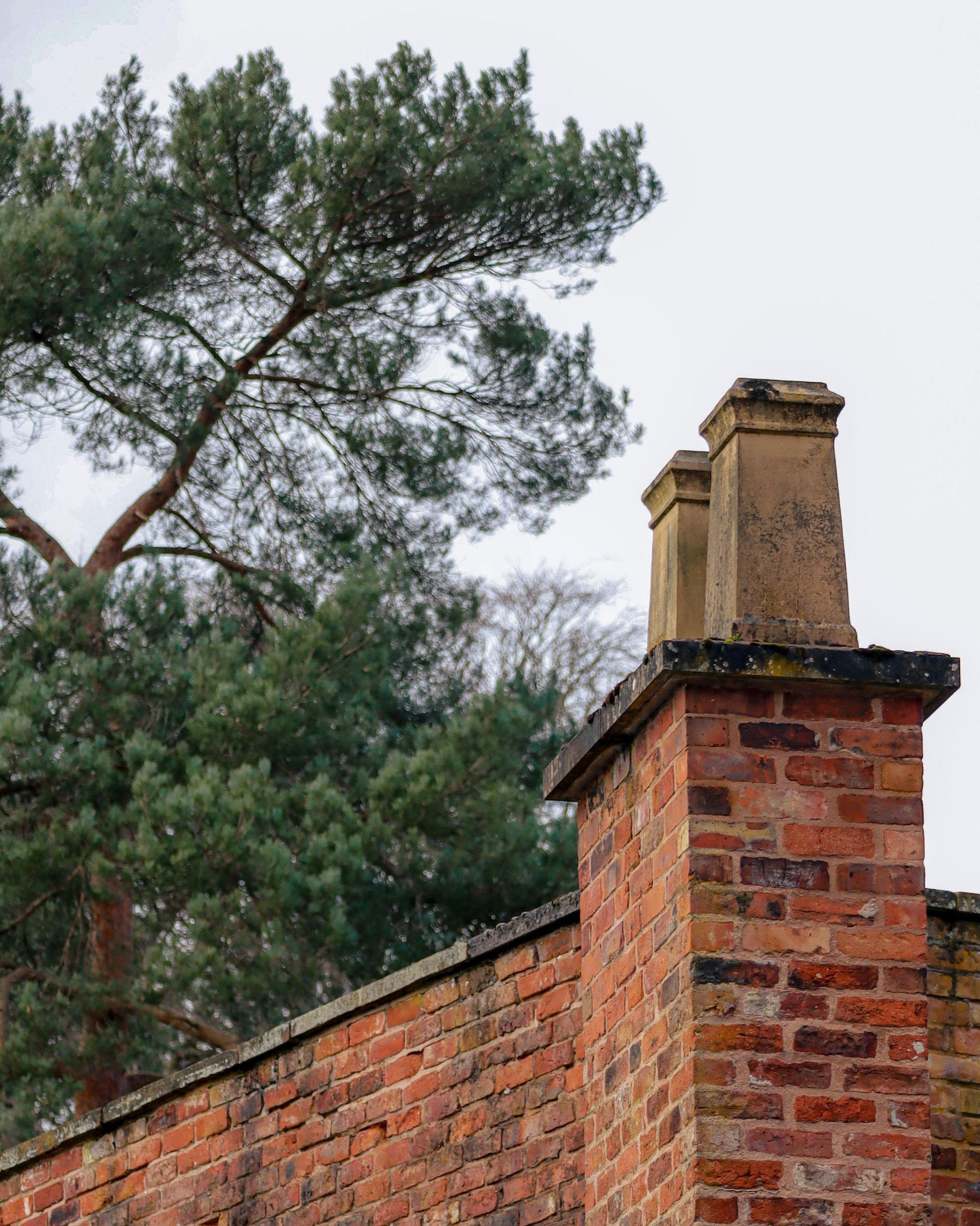 Brick chimney beside a red brick wall, with tall pine trees against an overcast sky