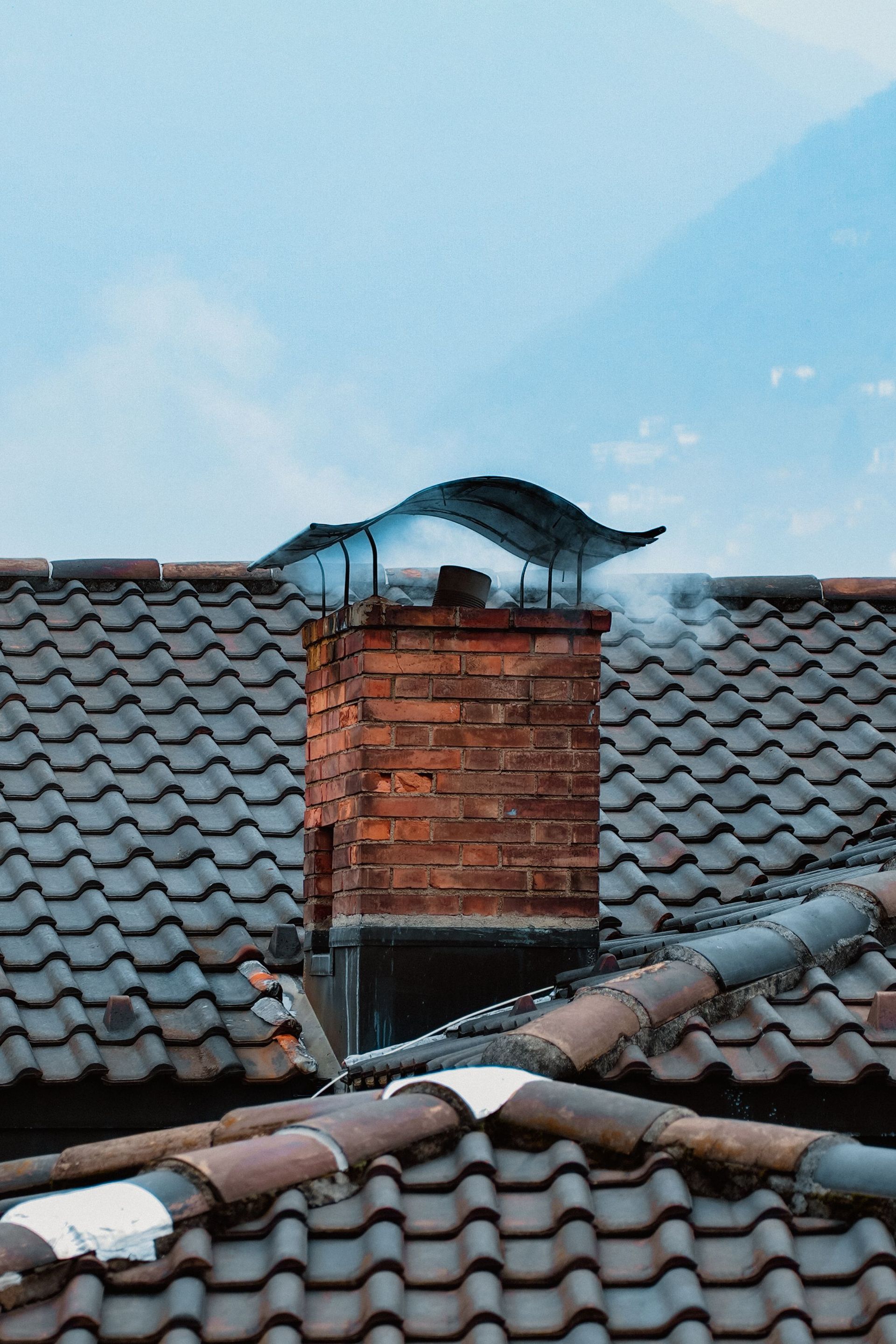Brick chimney on a tiled roof with smoke rising under a blue sky
