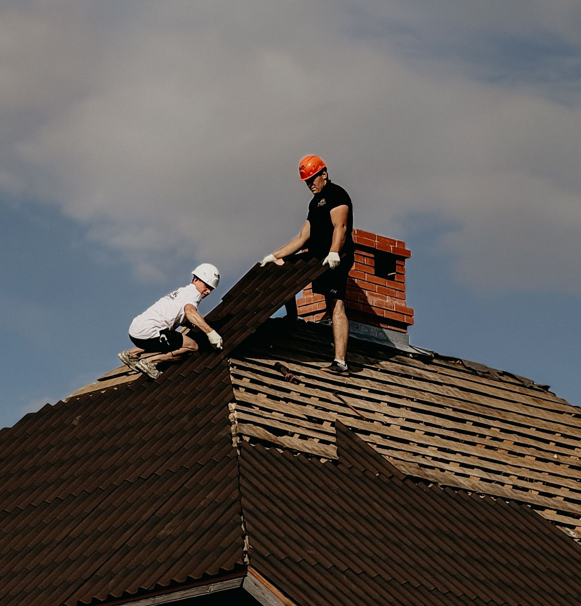 Two roofers replacing shingles on a house roof, one in a white shirt and one in black with an orange helmet.