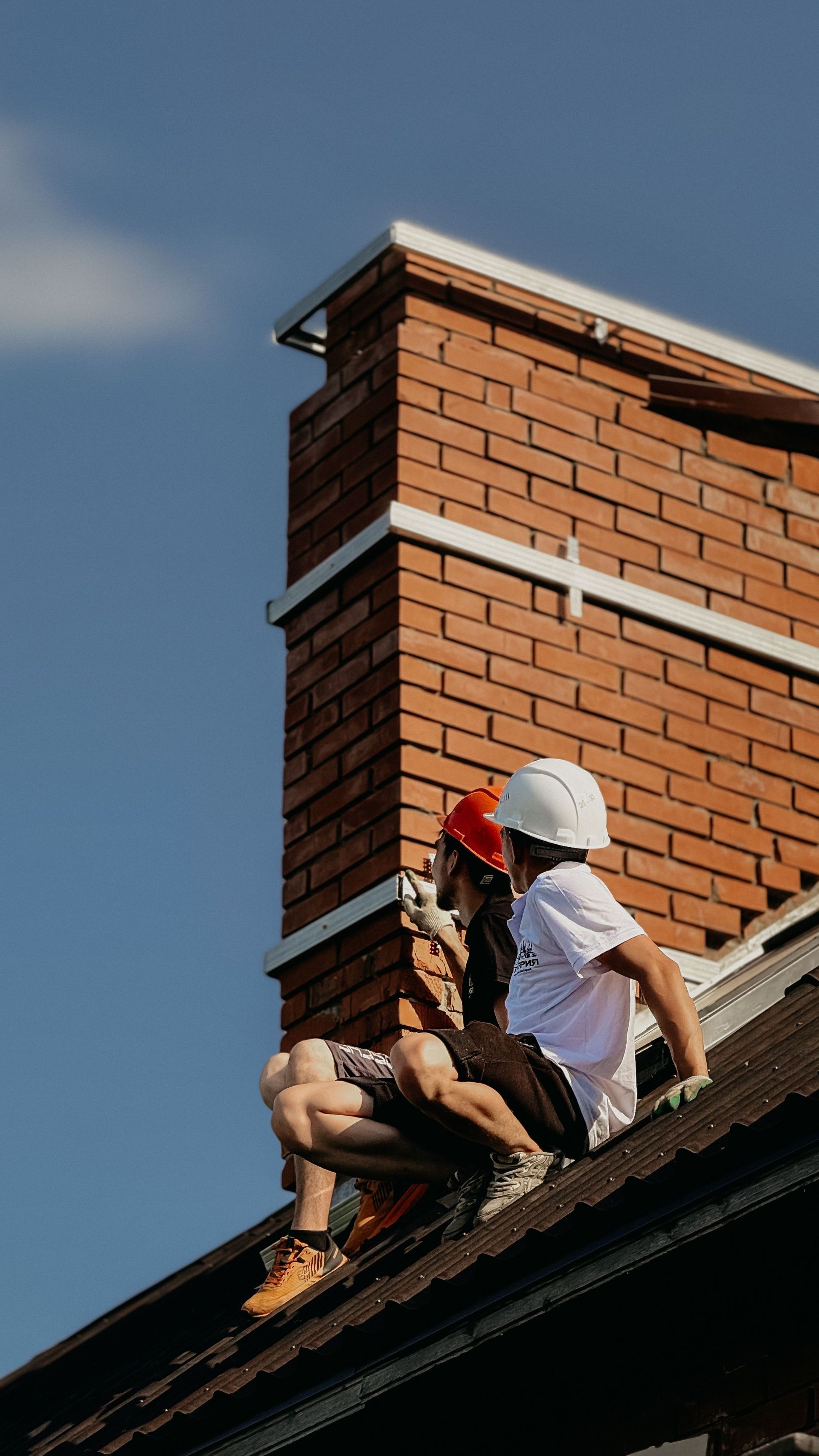 Two roofers working on a brick chimney, one using a power tool on a shingled roof.