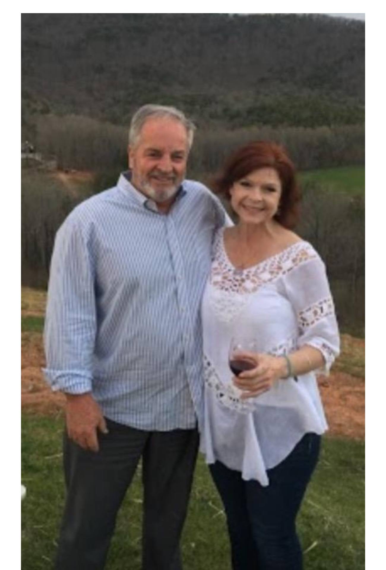 A man and a woman are standing next to each other in a field . the woman is holding a glass of wine.