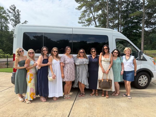 Group of women posing in front of a silver shuttle van outdoors on a sunny day