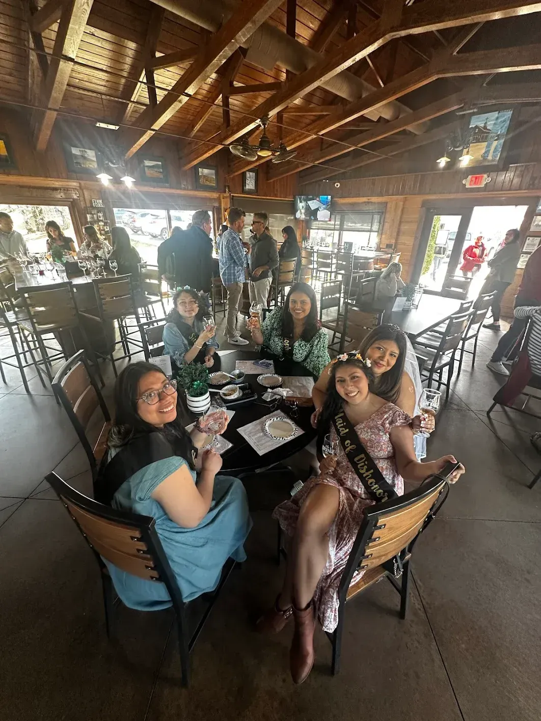 Group of friends at a restaurant, celebrating a woman with a sash, smiling, seated around a table.