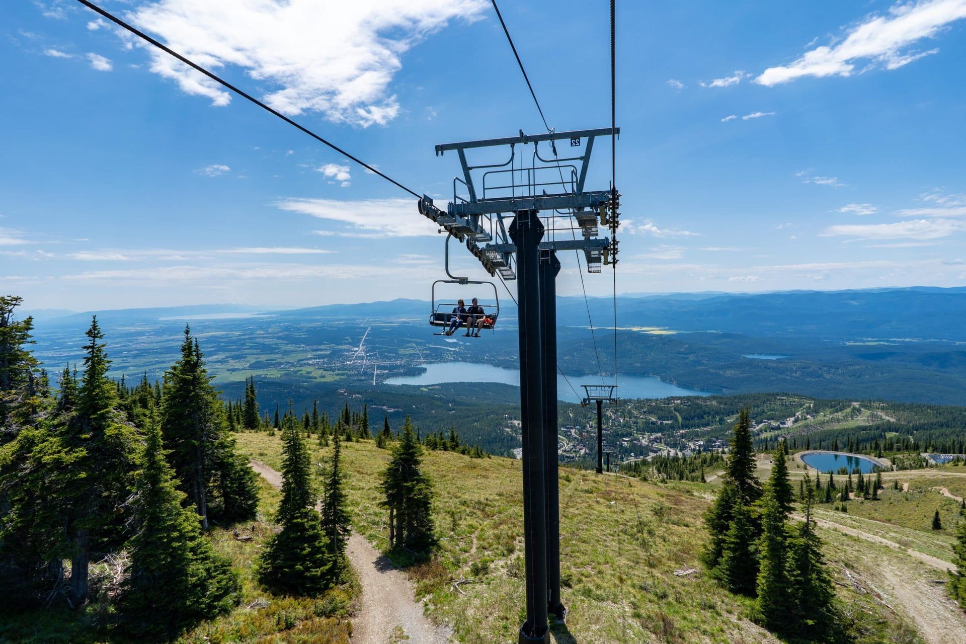 A ski lift with two riders ascends a mountain. Blue sky overhead, with trees and a lake in the distance.