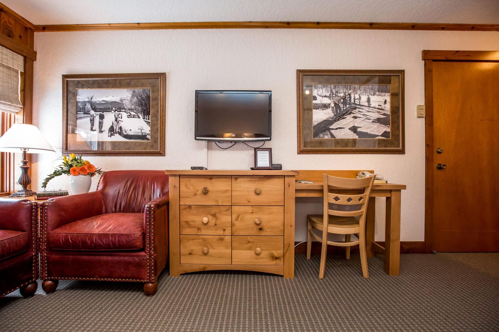 Hotel room with red leather armchair, wooden dresser, TV, desk, artwork, and closed wooden door.