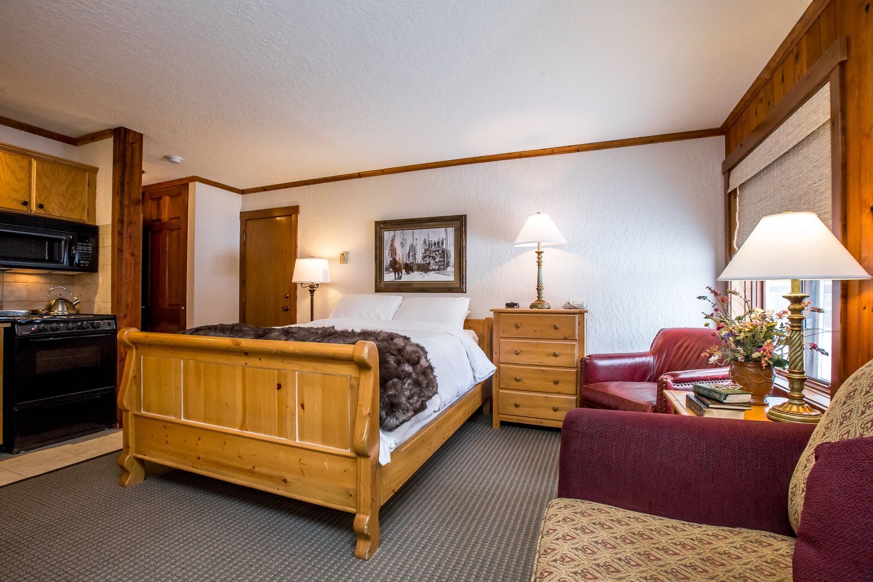 Cozy hotel room with wooden furniture. Bed, sofa, lamp, small kitchen. White walls, brown trim.