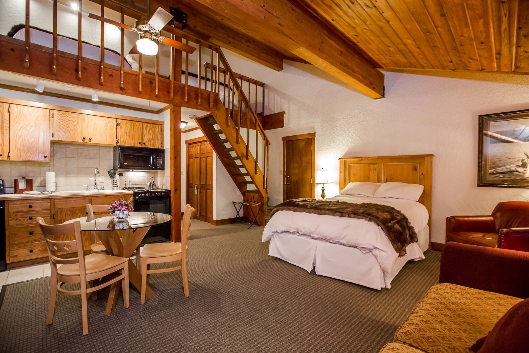 Cozy cabin interior with a loft, kitchen, dining area, and a bed in a room with brown wooden paneling and a ceiling.