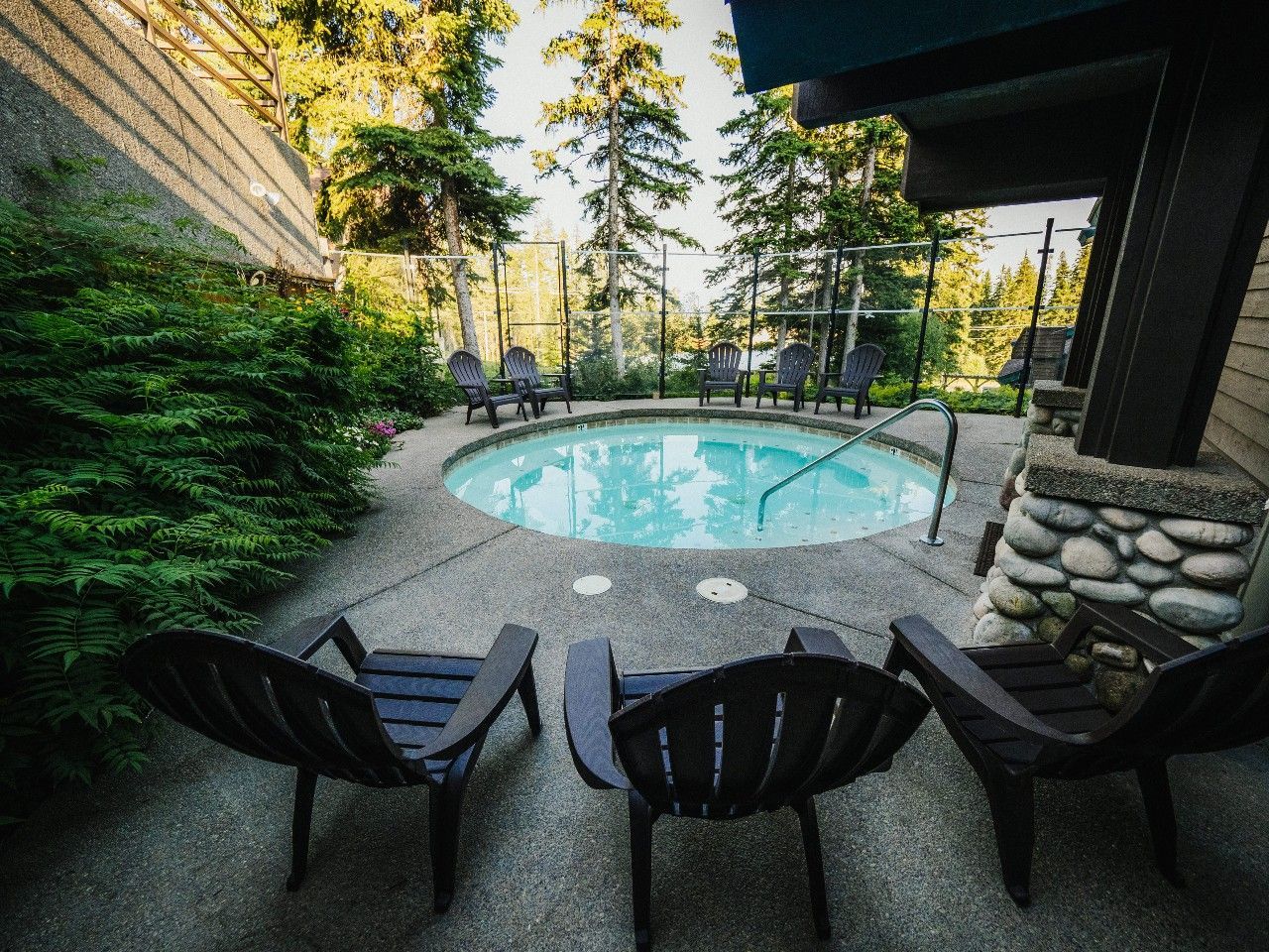 A circular outdoor hot tub surrounded by lounge chairs, set on a patio next to a wooded area.