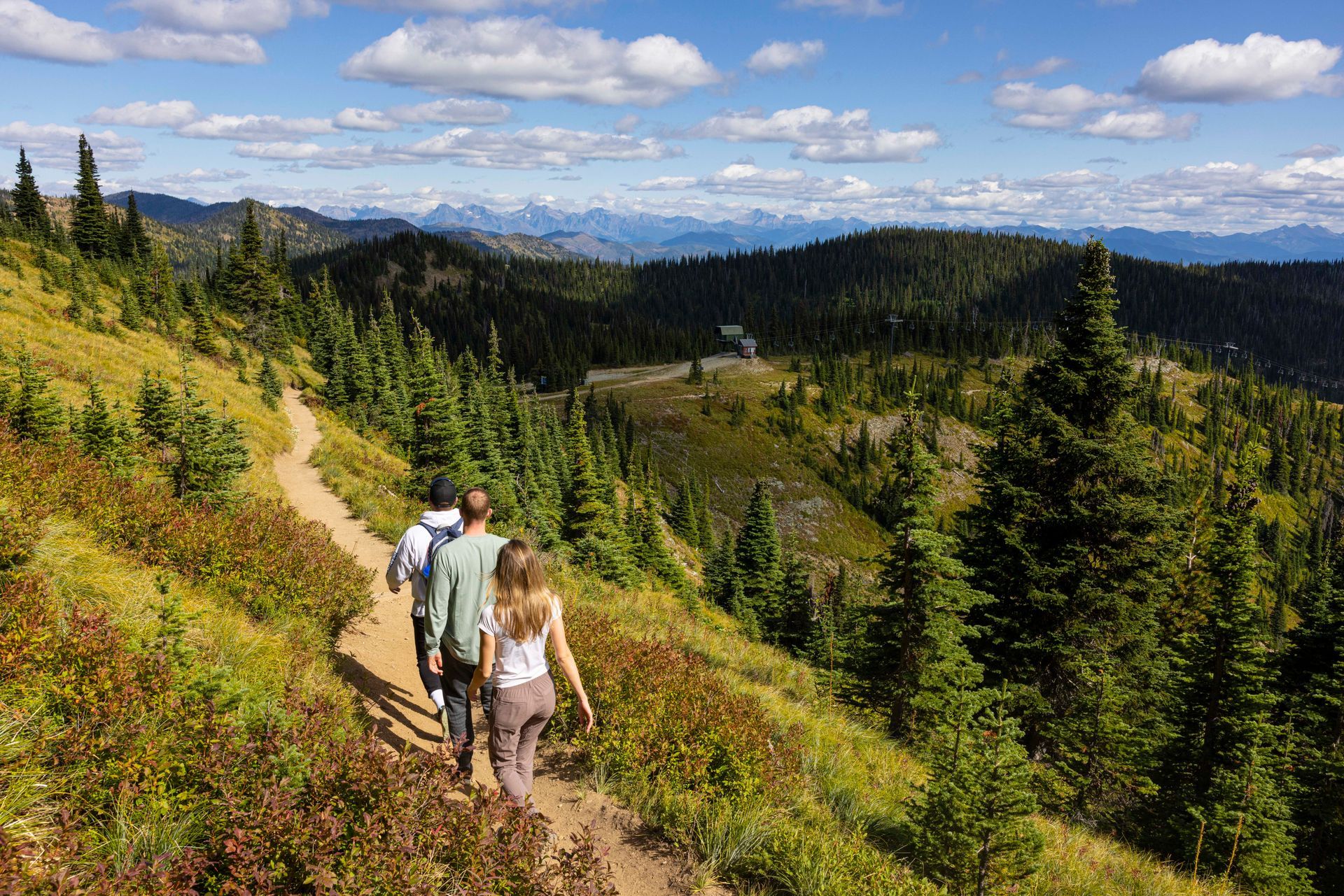 Three people hiking on a mountain trail, with a forested landscape and distant mountains under a blue sky.