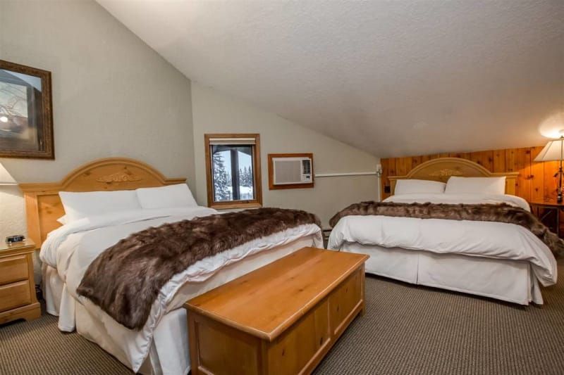 Two beds in a cozy room with wooden headboards and a storage chest in front. The room has a sloped ceiling and warm lighting.