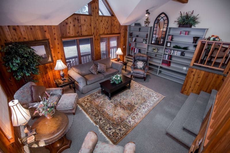 Cozy living room with wood-paneled walls, featuring a sofa, armchair, rug, bookshelves, and a small staircase leading up.