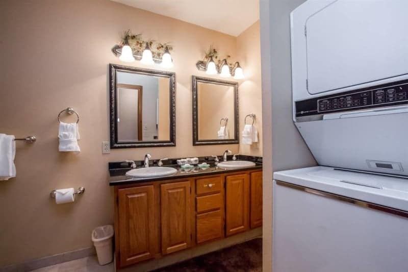 Bathroom with two sinks, brown cabinets, and a stacked washer and dryer. Beige walls and framed mirrors.