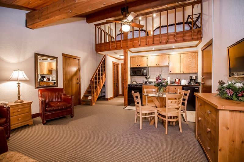 Cozy cabin interior with a loft bedroom, kitchen, dining area, and sitting area. Features exposed beams, wood furniture, and a staircase.