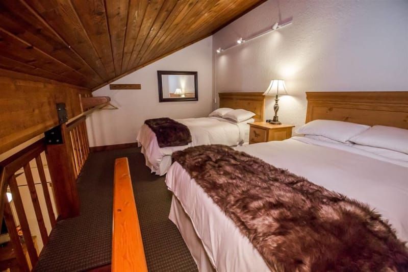 Loft bedroom with two beds, a double and a single, wooden ceiling and railing, fur throws, and a small bedside table.